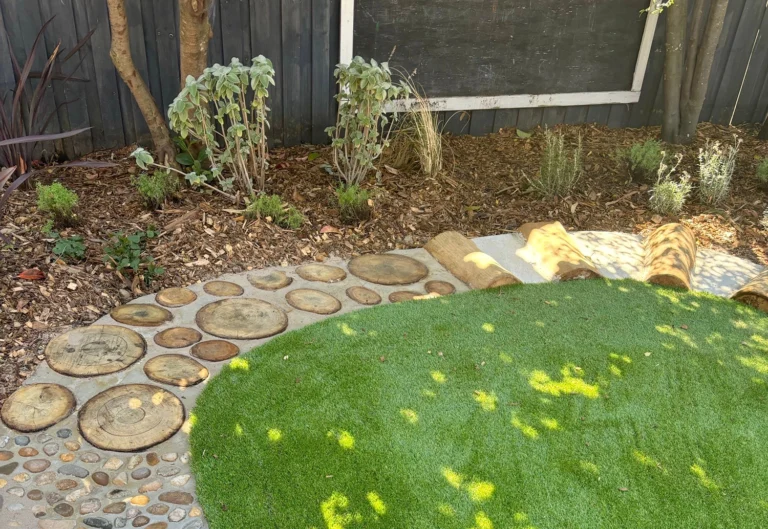 A backyard garden features a path made of circular wooden slices and small stones next to an area of artificial grass, with plants and shrubs in a mulched bed along the fence.