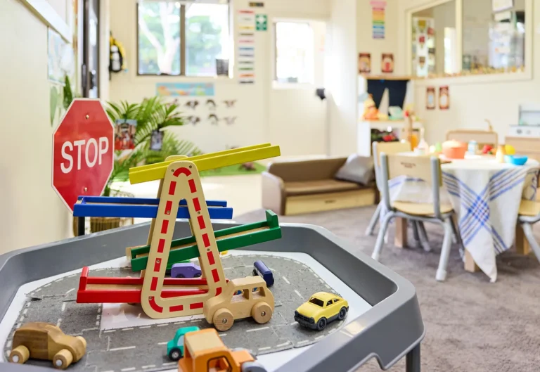 A well-lit room in a kindergarten with a children's play area featuring toy cars, a stop sign, and a ramp on a table. In the background, a table, chairs, couch, and educational posters are visible.