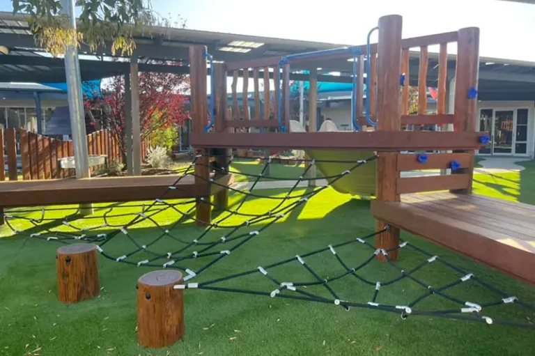 A playground structure featuring wooden platforms, rope climbing nets, and a slide, set on artificial grass under a canopy.
