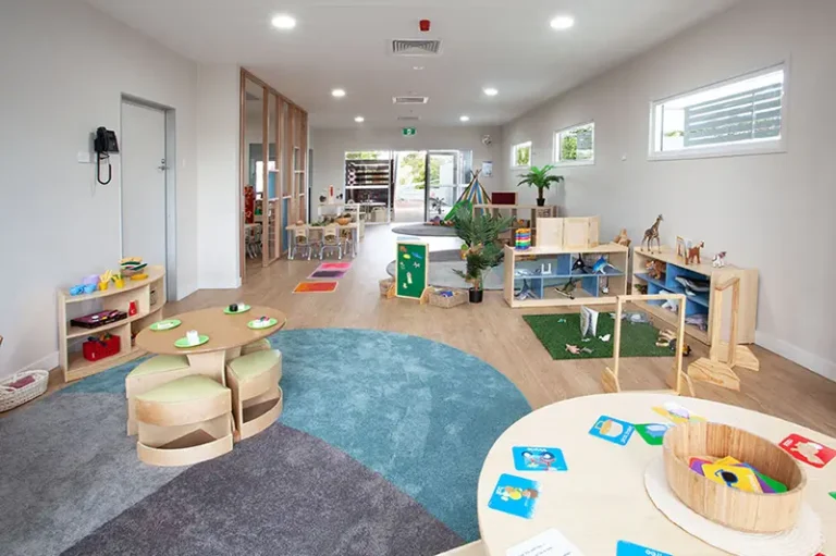A brightly lit, organized preschool classroom with circular wooden tables, chairs, educational toys, and books arranged neatly on shelves around the room. The flooring is a mix of blue carpet and wood.