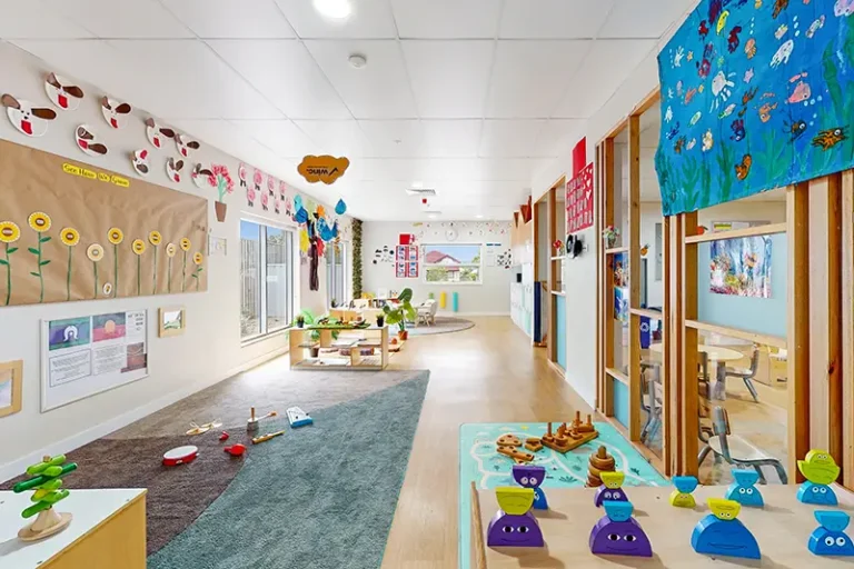 A brightly decorated, spacious classroom with art displays, wooden toys, and learning materials scattered around. Tables and chairs are arranged for different activities.
