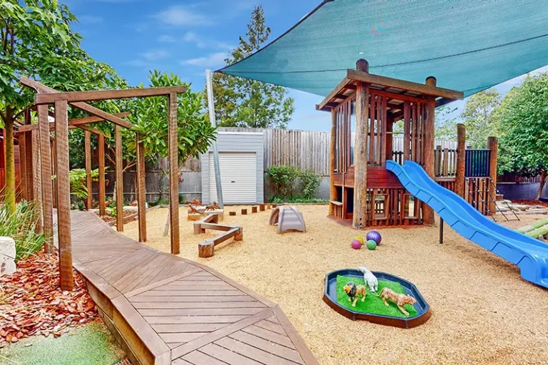 A playground features a wooden climbing structure, blue slide, shaded area, sandbox, and various play equipment set on a gravel surface with trees and fencing in the background.