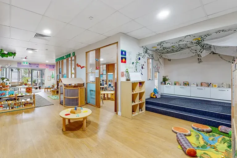 A brightly lit preschool classroom with shelves of toys, books, and educational materials. The room has wooden floors, a play area, and child-sized furniture.