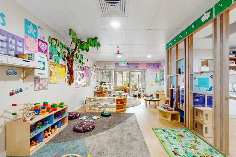 A brightly lit preschool classroom with various educational toys, colorful wall decorations, and activity stations. The room features a tree mural, play areas, and child-sized furniture.