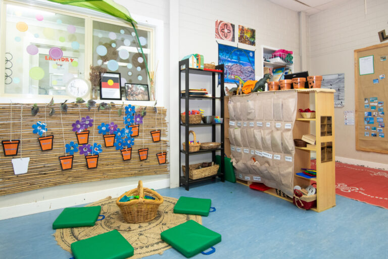 A colorful classroom corner tailored for early learning boasts a storage shelf, hanging wall organizers, vibrant green seating cushions, and a playful area rug. The walls are adorned with engaging posters, crafts, and charming potted plant decorations.