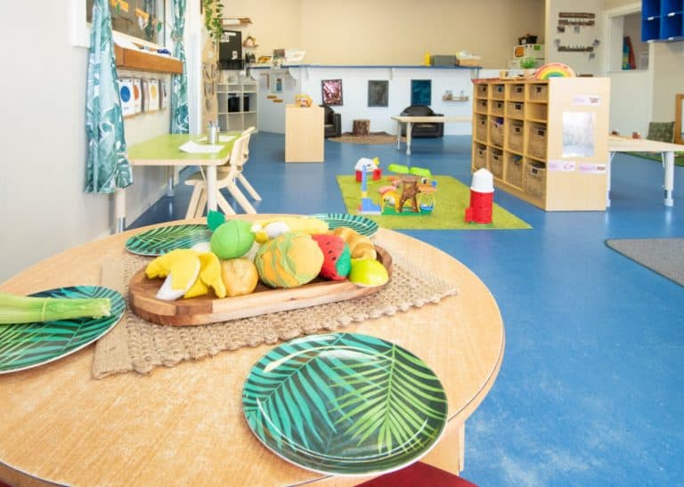 A bright, organized preschool classroom with a table set with toy fruits and green leafy plates, shelves with various toys, and a play area featuring a small wooden structure on a green mat provides an ideal early learning environment.