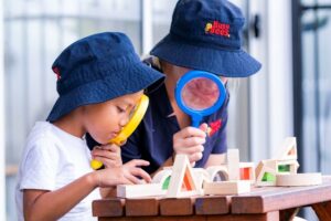 Two preschool children in hats use magnifying glasses to examine wooden block structures on a table, fostering early learning and curiosity.