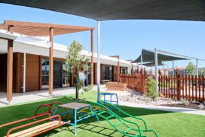 A modern playground with colorful climbing structures and artificial grass, perfect for preschool adventures. Wooden buildings and shaded areas in the background under a clear blue sky create an inviting space for early learning and day care activities.