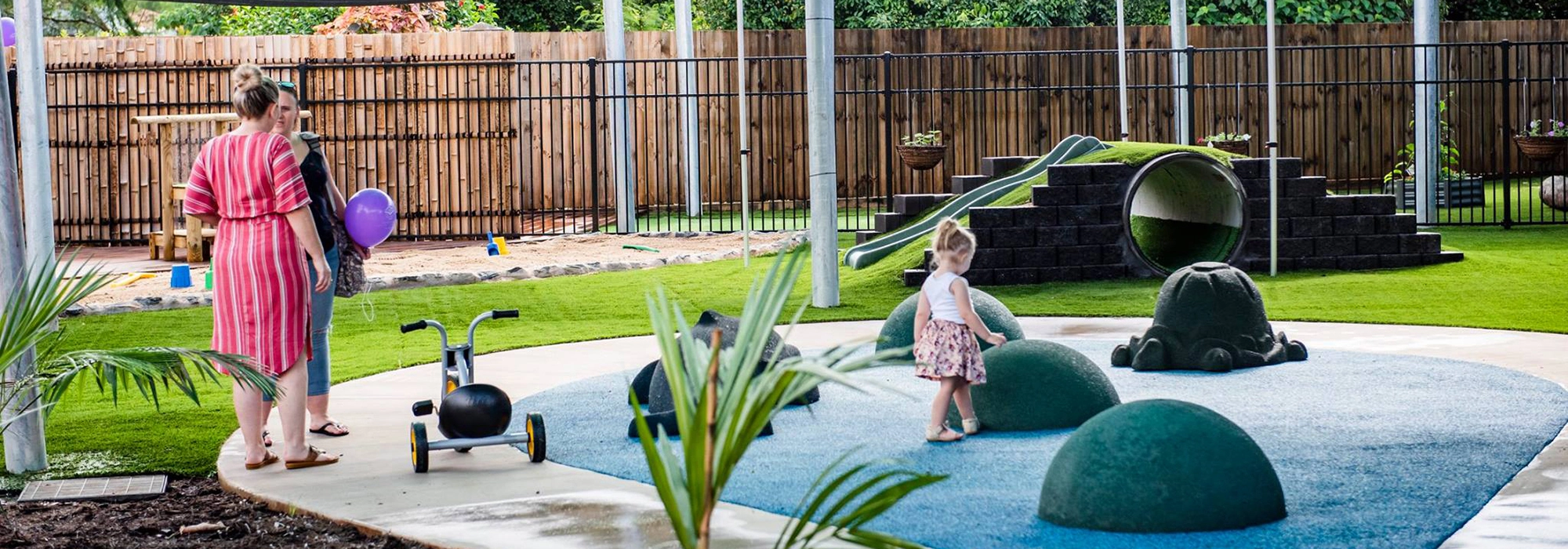 A woman and child stand near a tricycle while a little girl plays on dome-shaped structures at an outdoor playground. The area has artificial grass, a slide, and is fenced with wooden panels.