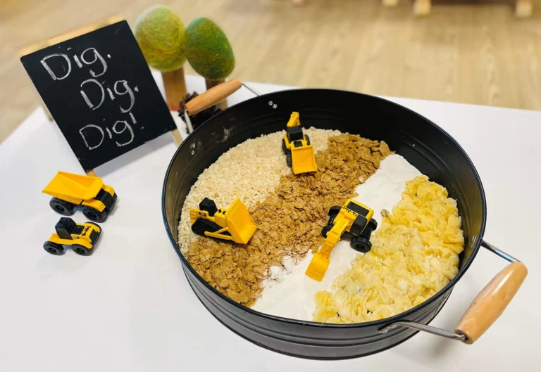 A round black tray contains toy trucks on sections of rice, oats, flour, and pasta. Nearby, two toy trees and a small sign with "Dig Dig Dig!" written on it accompany two additional toy trucks.