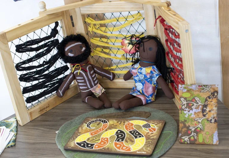 Two culturally themed dolls with dark hair and colorful clothes sit on a wooden surface. Behind them is a wooden frame with looped strings, and a colorful, patterned board is in front of them.