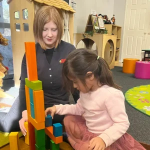 A woman and a young girl are sitting on the floor of a bright preschool, playing with colorful building blocks amidst children's play equipment and toys.