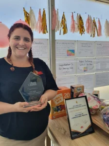 A woman holds a trophy and stands beside an award certificate on a table, with party decorations and congratulatory notes from her preschool displayed on the wall behind her.