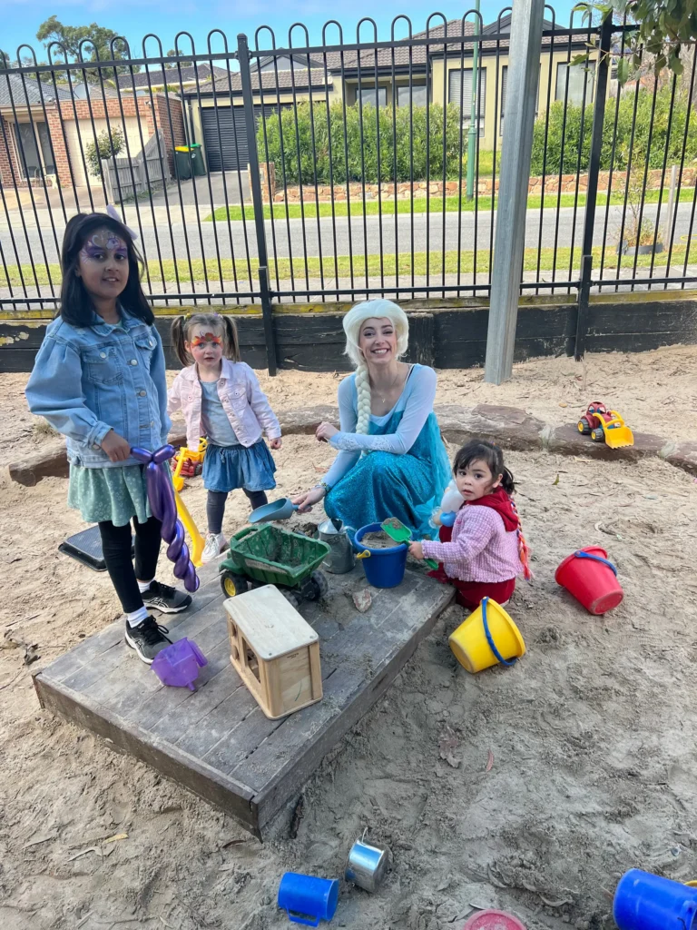 Four children play together in a sandbox with various toys like buckets and trucks, in front of a black fence and a building in the background. Three are standing, and one is sitting, enjoying their time in this vibrant kindergarten setting.