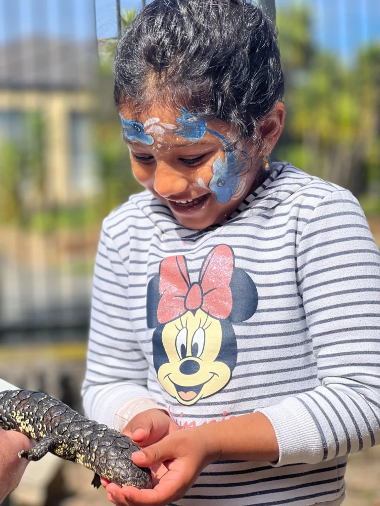 A child from daycare with face paint and wearing a striped Minnie Mouse shirt joyfully holds a lizard and smiles.