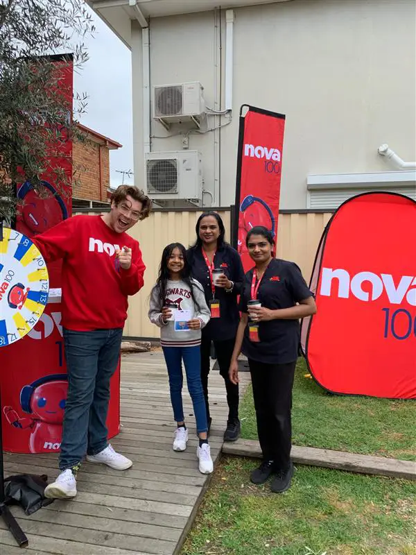 Four people stand outside next to a "Nova 100" banner and a prize wheel. One person holds a branded cup and another holds a drink. They all appear to be engaging with the event, much like teachers at an early learning center fostering community spirit.