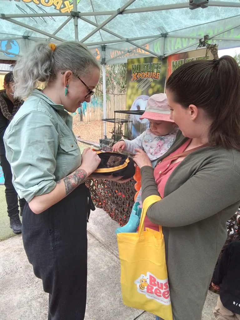 A woman holds a young child as another woman shows them a small animal at an outdoor kindergarten event. The young child reaches out towards the animal.