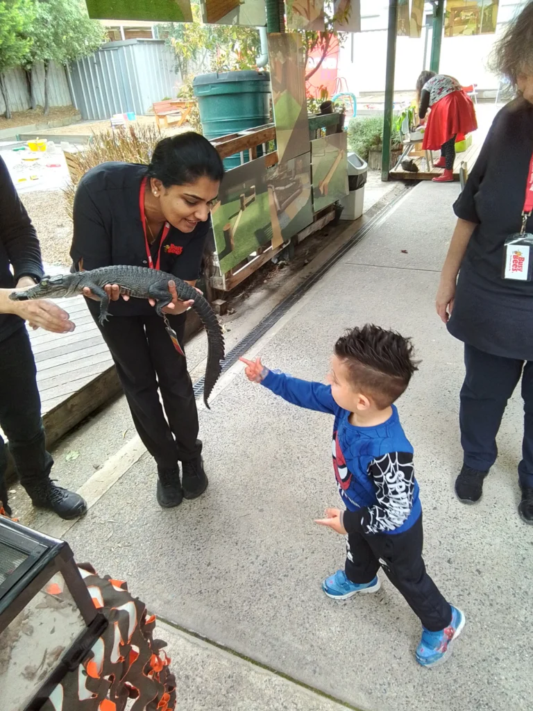 A woman holding a small alligator leans down to show it to a young child dressed in a Spiderman outfit, who is pointing at the alligator. Other adults are partially visible in the background, creating an engaging scene that could easily be from a day care or early learning environment.