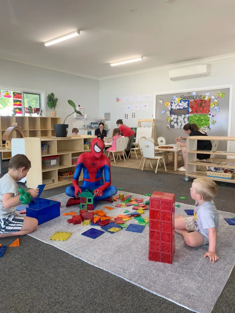 Children play with colorful building blocks in a day care classroom. An adult dressed in a Spider-Man costume sits among them, also engaged in building with blocks, creating a fun and imaginative environment for early learning.