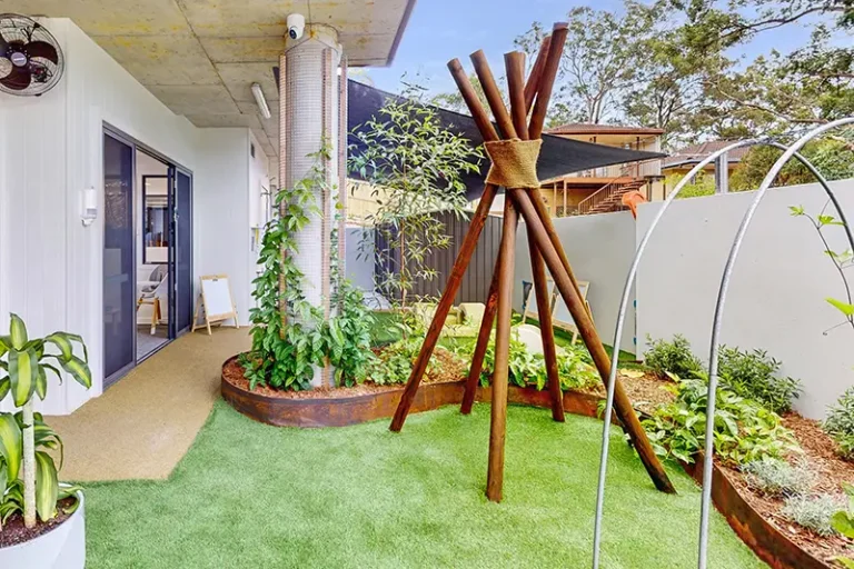 A small balcony garden with green plants, raised garden beds, and a wooden teepee structure. The balcony is adjacent to a sliding glass door leading into a modern apartment, perfect for families prioritizing early learning. Trees are visible in the background, adding to the serene atmosphere.