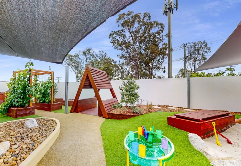 A playground features a wooden climbing structure, raised garden beds, a sandpit with toys, and a water play station, all under shade sails with trees and a blue sky in the background.