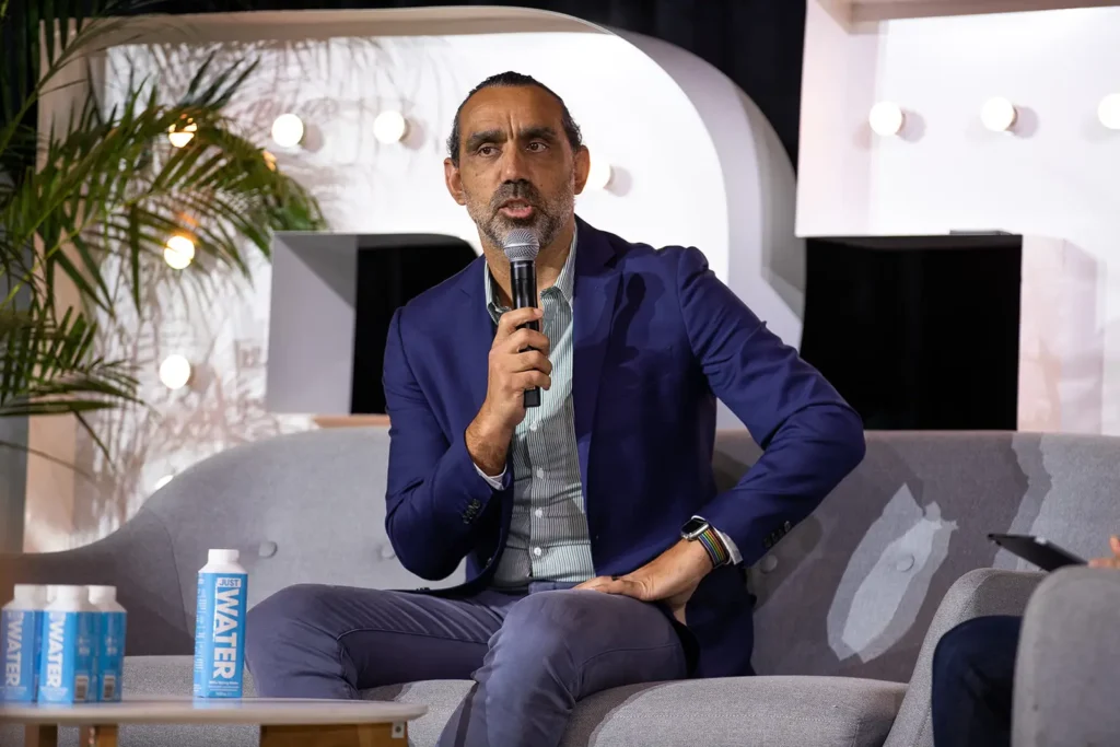 A man with a beard and wearing a blue jacket speaks into a microphone while sitting on a grey couch, discussing the importance of early learning. There are water bottles on a table next to him and plants in the background.