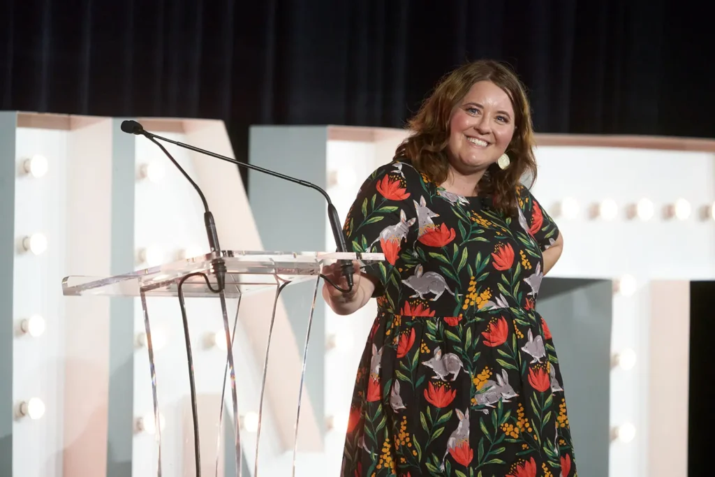 A woman in a floral dress stands by a clear podium with a microphone, smiling, with large illuminated letters in the background, celebrating the grand opening of a new kindergarten.