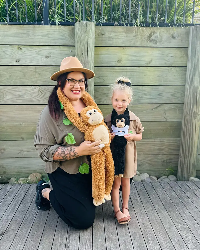 An adult and a child smile at the camera while holding plush animal toys, standing on a wooden deck with a wooden fence in the background. The child, proudly displaying a certificate, celebrates an achievement from their preschool early learning program.