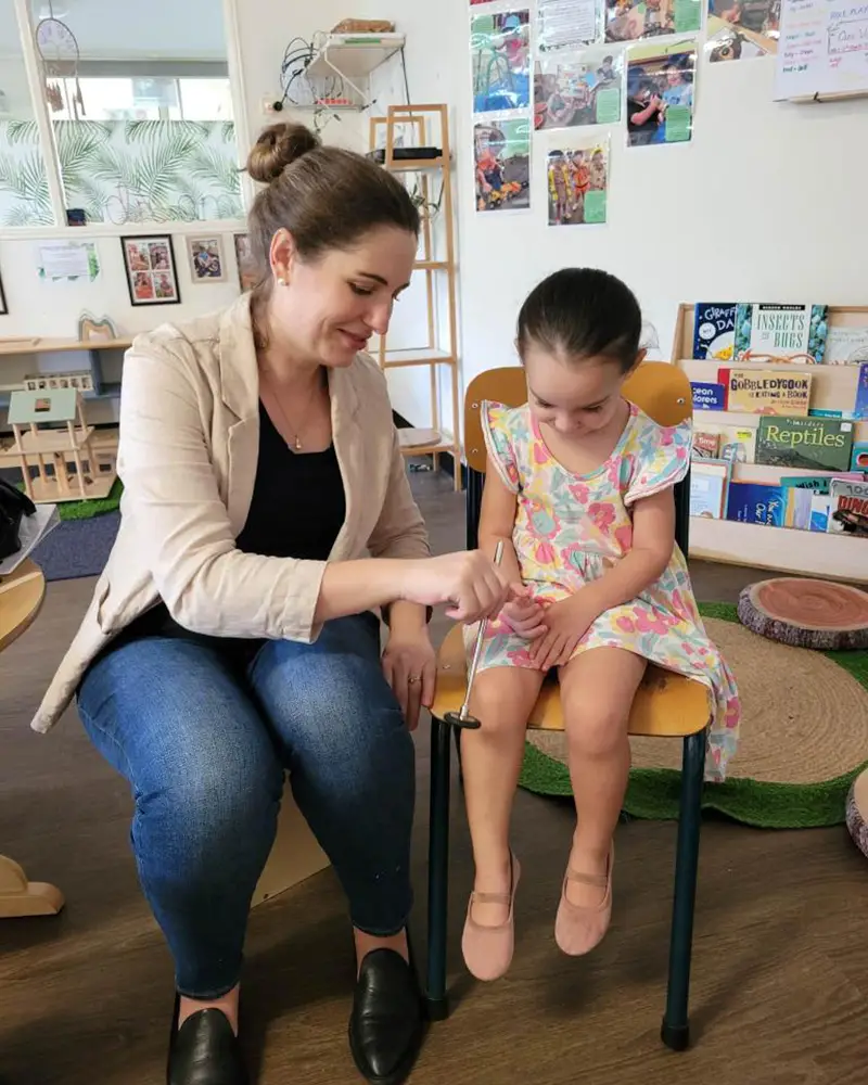 A woman is sitting next to a young girl who is seated on a chair. The woman is holding a stethoscope to the girl's knee. They appear to be in a kindergarten classroom or play area with books and decorations in the background, fostering an environment of early learning.