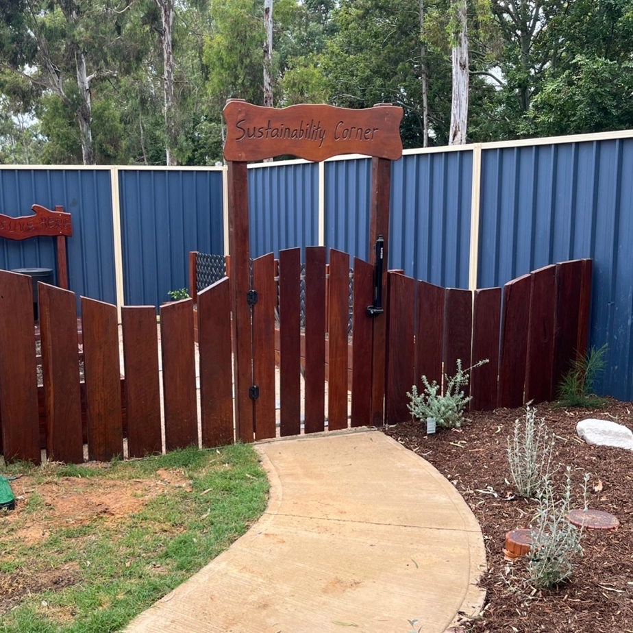 A wooden gate labeled "Sustainability Corner" opens to an enclosed area with blue metal fencing, surrounded by trees. A curved concrete pathway leads to the gate, and plants line the path.