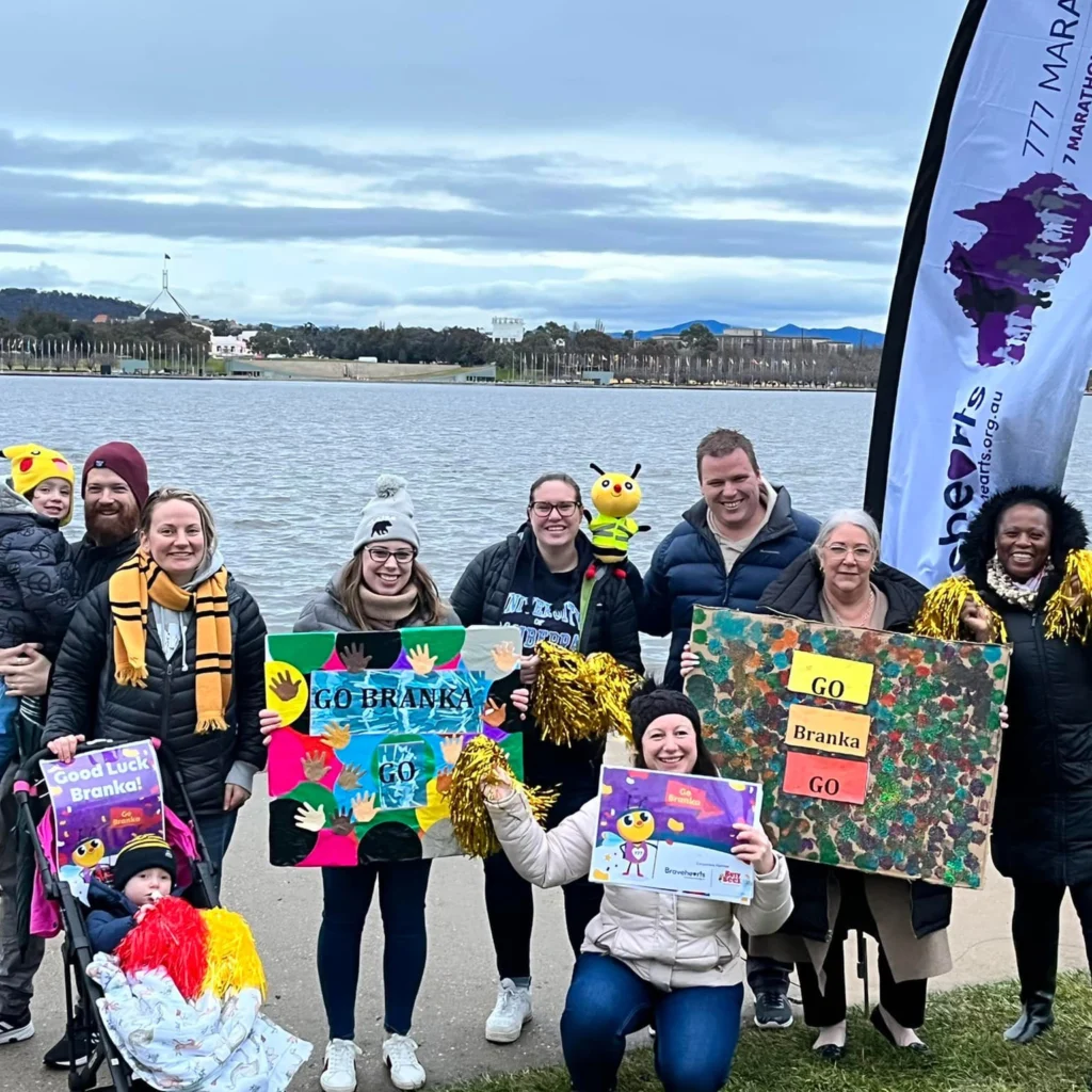 A group of people stand outdoors holding signs of support and smiling as they pose for a photo. Some are wearing hats and scarves, and a baby is in a stroller. The background includes a lake and overcast sky.