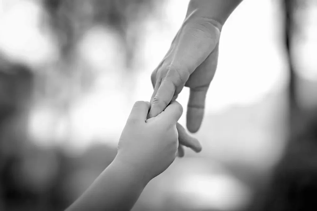 A close-up image in black and white showing an adult hand gently holding a child's hand outdoors.