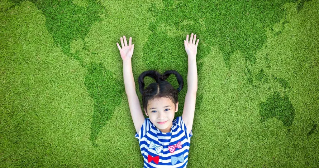 A young girl lies on green grass with her arms extended above her head, forming the shape of a map of the world. She's wearing a striped shirt and smiling.