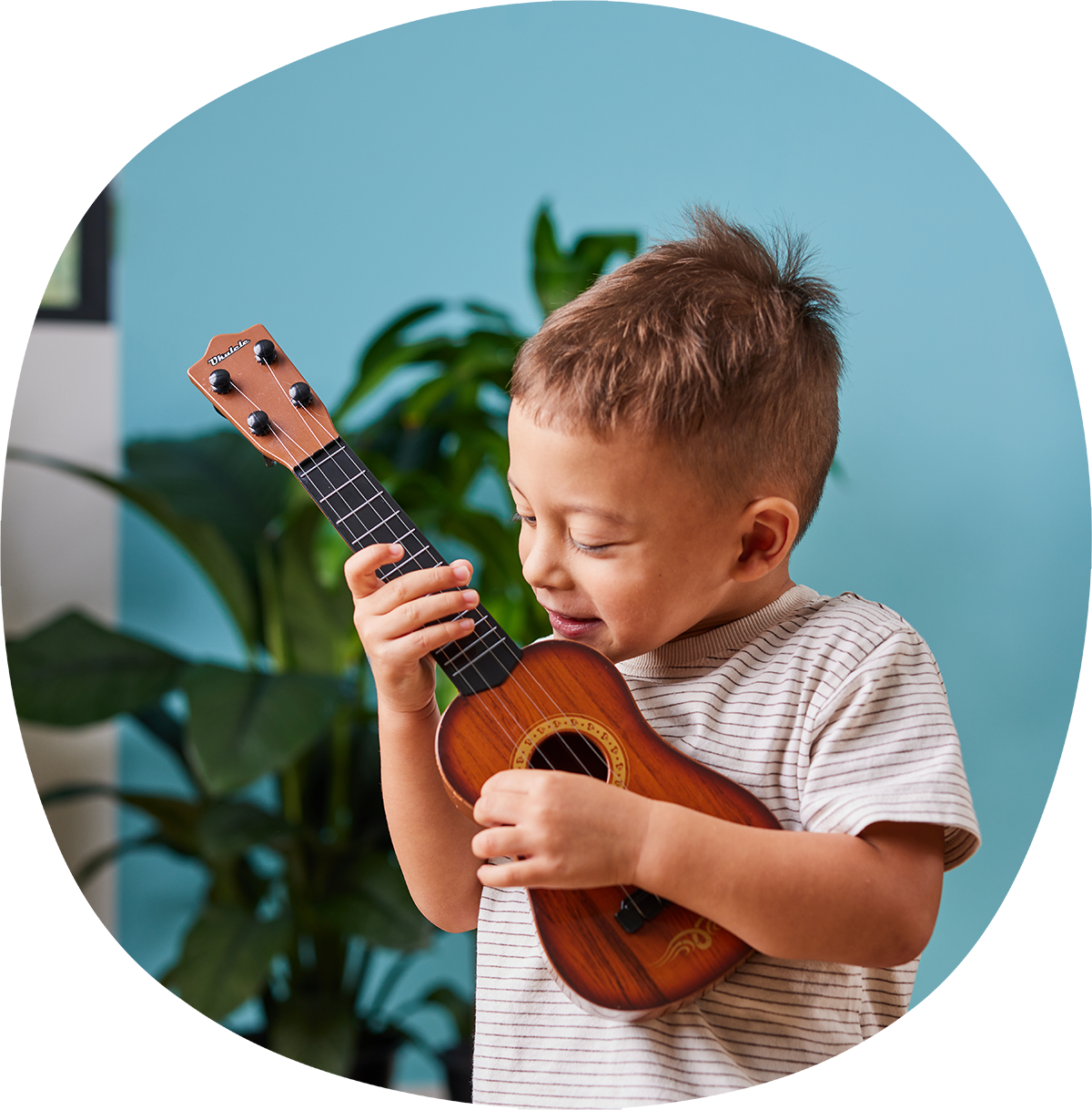 A young boy with short hair is playing a small wooden ukulele. He is wearing a striped t-shirt and standing in front of a blue wall with green plants in the background.