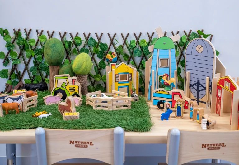 A tabletop farm playset with wooden animals, structures, and a toy tractor, set against a green backdrop with artificial grass and foliage.