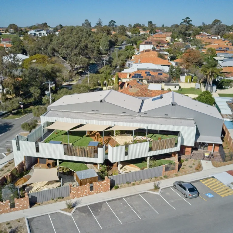 Aerial view of a childcare center with a rooftop playground, adjacent parking lot, and surrounding suburban neighborhood with trees and houses.