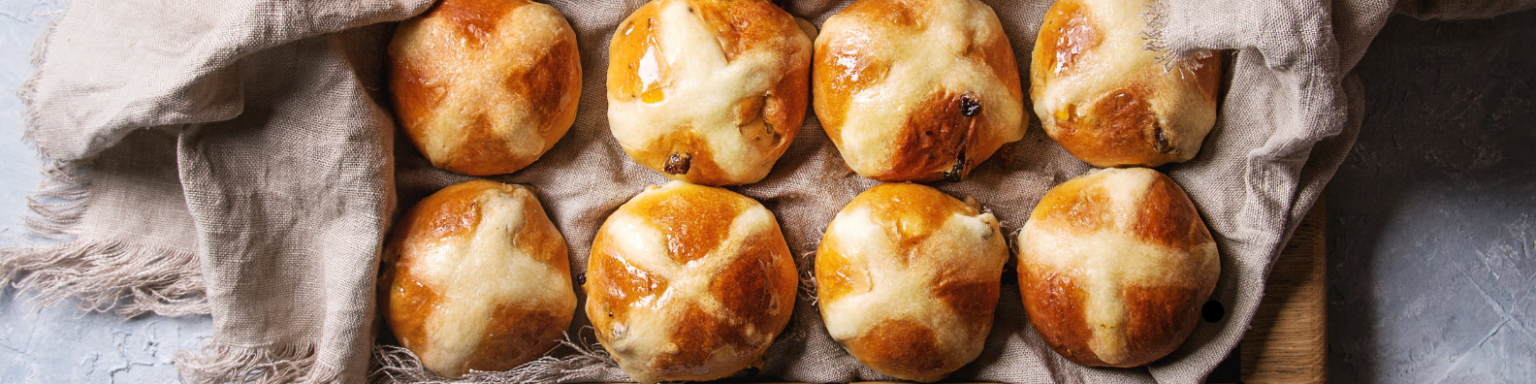 Eight glazed hot cross buns on a textured cloth, arranged in two rows on a gray surface.