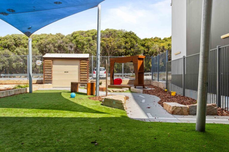 Outdoor play lawn at Busy Bees Aveley with synthetic grass, a timber gate and bushland beyond