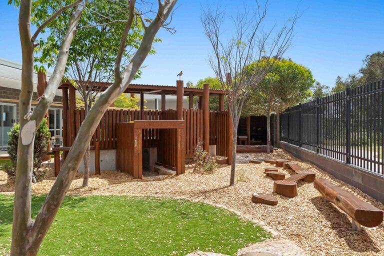 Timber play fort at Busy Bees Banksia Grove Village with gum trees, log stepping stones and synthetic grass