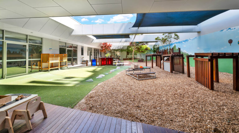 Covered outdoor courtyard at Busy Bees Glenroy Station with synthetic grass, bark softfall and a sky mural
