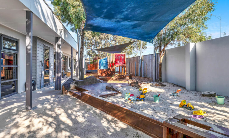 Large shaded sandpit at Busy Bees Truganina with a timber deck, natural rocks and sand toys