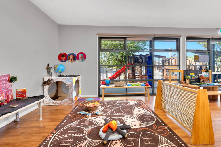 Indoor storytelling space at Busy Bees Truganina with a pretend campfire on a patterned rug and a view to the playground